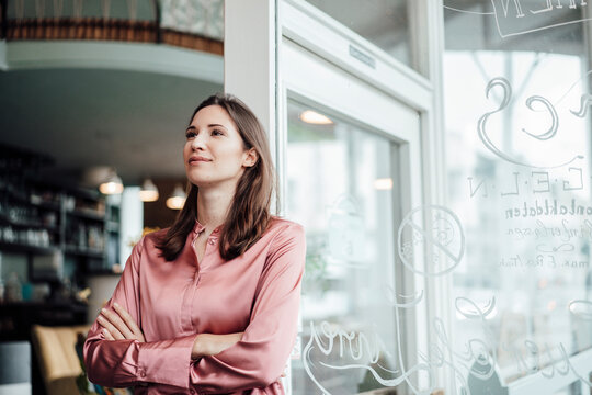 Female entrepreneur with arms crossed leaning against glass wall in cafe