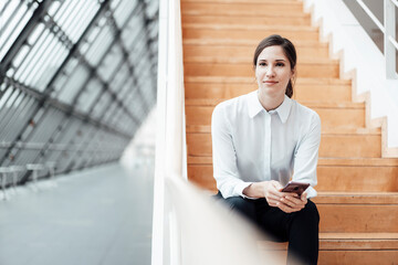 Smiling businesswoman with smart phone sitting on steps in corridor
