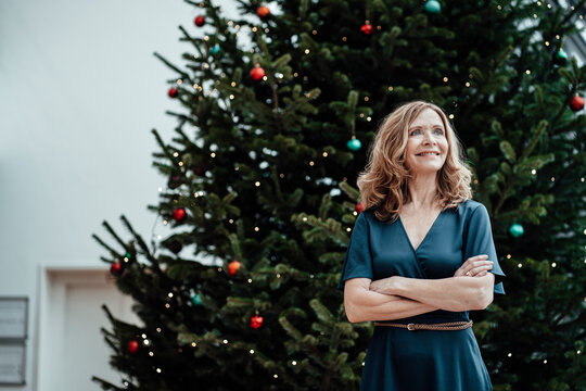 Smiling businesswoman with arms crossed looking away against Christmas tree in office - Powered by Adobe