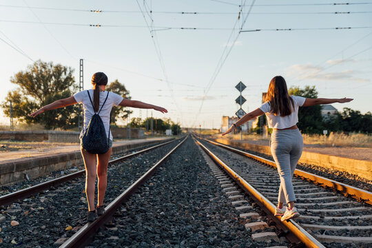 Female friends with arms outstretched walking on railroad track against sky at sunset