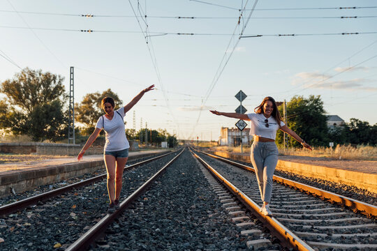 Carefree female friends with arms outstretched walking on railroad track against sky at sunset