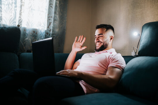 Young businessman waving hand on video call through laptop while sitting on couch in living room