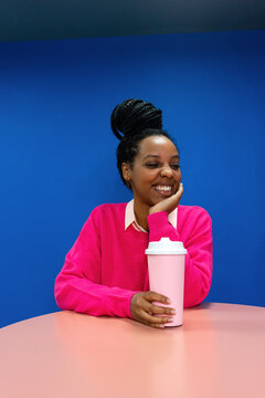 Smiling young businesswoman with hand on chin holding reusable coffee cup against blue wall in office
