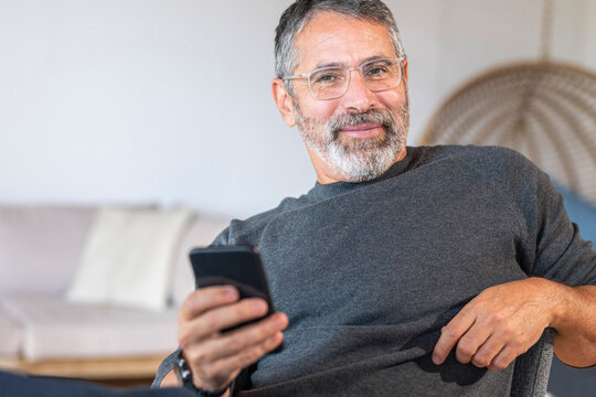 Smiling businessman wearing eyeglasses using mobile phone while sitting at home