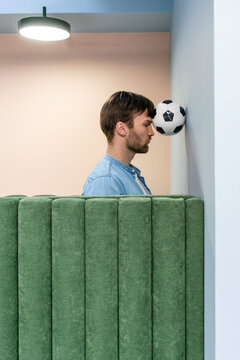Male entrepreneur pressing soccer ball with head on wall at workplace