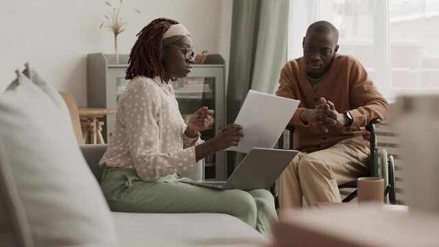 Medium shot of young African businesswoman holding documents in hands while sitting on sofa in coworking space and discussing them with disabled colleague sitting nearby on wheelchair