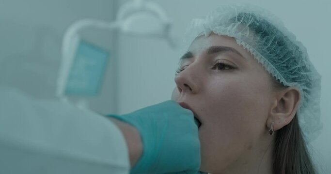 Close-up a young female patient in disposable hat, being examination a mandibular by dentist using his fingers while she is opening her mouth.