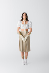 full length of brunette woman in gloves with flowers and pleated skirt standing on white