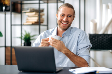 Fototapeta na wymiar Happy friendly confident caucasian guy, employee or freelancer, holding a cup of coffee while sitting at his desk at home or office, looking at the camera with a friendly smile