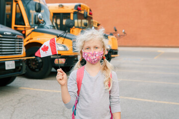 Caucasian girl student in protective face mask with Canadian flag. Student kid near yellow school bus outdoor. Education and back to school. New normal during coronavirus covid-19.