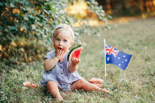 Cute adorable Caucasian baby girl eating ripe watermelon with Australian flag on ground. Funny child kid sitting in park with fresh fruit celebrating Australia Day holiday outdoor.