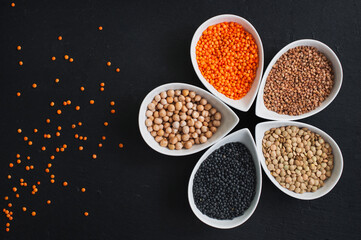Different types of lentils in ceramic bowls (red turkish lentils, canadian lentils, black lentils - beluga, buckwheat, chickpeas) on black slate table.. For culinary, shop, restaurant. Top view .