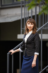 Fototapeta premium Young woman in classic business suit on background of a City park. Business style girl in Summer City on background of new park near a building. Street style Modern strict urban girl.
