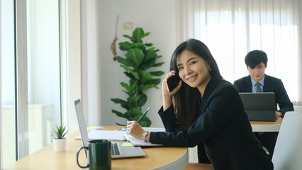 Happy businesswoman working with computer laptop and talking on mobile phone while sitting with her colleague in modern office.