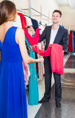 Young smiling man helping his girlfriend to choose gown in clothes store