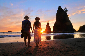Silhouette of couple on beach walking towards rising sun with sea stacks on background. Rialto...