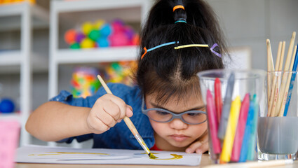 Young Asian down syndrome little girl sitting at desk with colors and painting with concentrated and calm. painting. Concept for education for disabled kid © Bangkok Click Studio