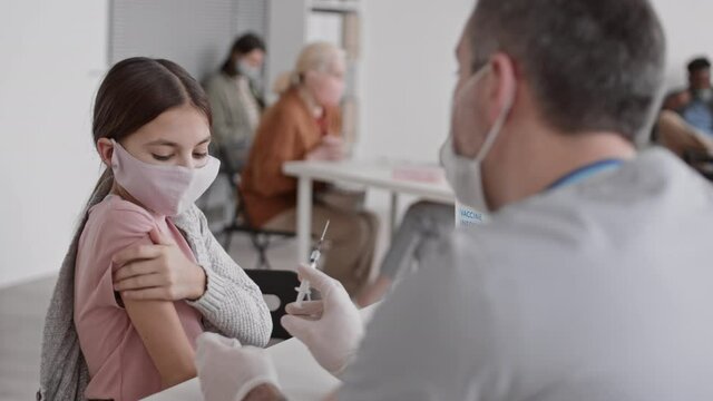 High angle medium close-up of school-aged Caucasian girl wearing protective face mask, sitting at table in clinic, vaccinated on shoulder by unrecognizable medical worker, then leaving