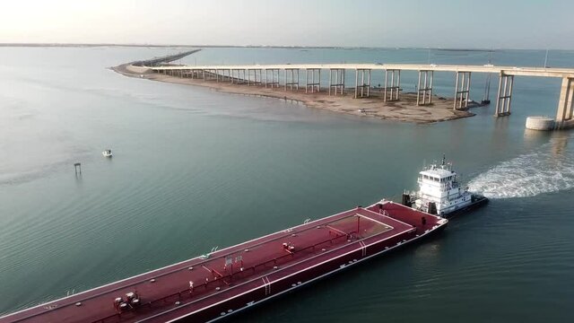 Rotating aerial view of large red barge maneuvering along the Gulf Intracoastal Waterway into Laguna Madre after passing under JFK Memorial Causeway Bridge near sunset; Corpus Christi Texas