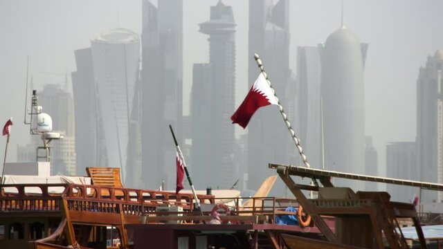 Flag Of Qatar Waves In Wind Above Wooden Dhow Boats And Before A Massive Cityscape Of Skyscrapers In Sunshine