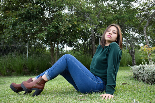 Mujer Joven Latina sentada en un jardín sobre grama verde, usando una blusa verde, pantalón azul y botas color café. Chica Guatemalteca muy linda.