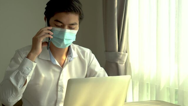 Asian man in protective facial mask taking a call on his mobile phone on his laptop with a serious concerned expression, Domestic violence, Stress, Liabilities