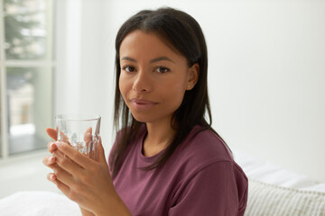 Portrait of cute mixed race girl in casual clothing relaxing in bedroom after awakening drinking...