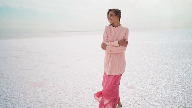 slow motion wonderful dreamy woman in big kozy sweater and pink dress standing at windy salty coastline on pink salt lake. inspired paradise picture, wellness and travel concept. nature in Ukraine