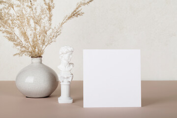 Blank paper card, invitation mockups with small statue and pampas grass in vase on white and beige background. Modern home decor concept. copy space