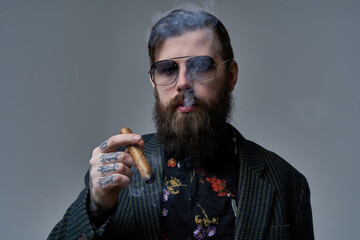 Portrait of a serious and handsome man with stylish coiffure posing in gray background. Tattooed man smoking cigar.