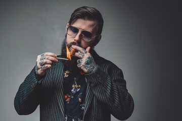 Tattooed macho smoking cigar and dressed in fashionable suit poses in gray background. Portrait of...