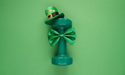 Heavy dumbbell wearing Irish shamrock leaf clover hat and a bow tie. Healthy fitness gym flat lay concept for St. Patrick's Day. Composition on green background.