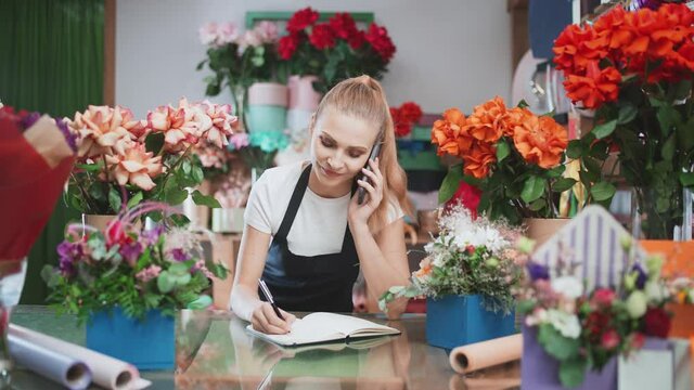 Florist female discusses the order by talking on the mobile phone and using a notepad in a flower shop.