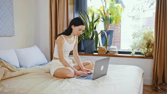 Freelance career. Young woman with vitiligo skin sitting on bed and working on laptop computer, tracking shot