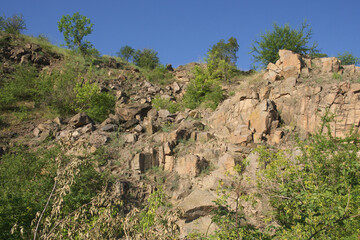 Fototapeta premium A rocky slope of an abandoned granite quarry in the Nikolaev region of Ukraine on a hot summer day. Sharp stones overgrown with sparse vegetation. Blue cloudless sky over the cliffs.