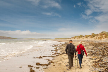 Senior couple walking on a sand by the ocean. Cloudy sky. Mature relationship and active outdoor activity for elder persons concept.