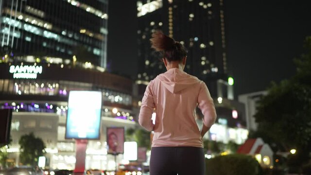 Slow-motion,Back Follow Camera View.Female athlete in hooded shirt Jogging at night City streets with lots of lights in the background.