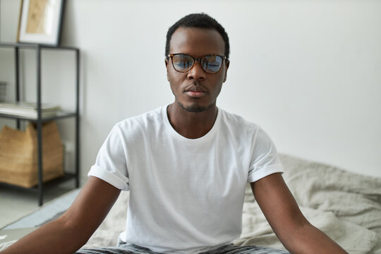 Self-conscious young dark skinned male in eyeglasses sitting on bed with eyes closed, having calm peaceful expression, focused on breathing, practicing mindfulness meditation in the morning