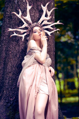 Portrait of Sensual Caucasian Girl Posing With Artistic Deer Horns and Long Light Dress In Summer Forest.