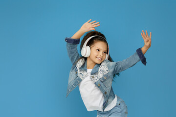 adorable smiling little child girl in white headphones listening to music and dancing on blue background