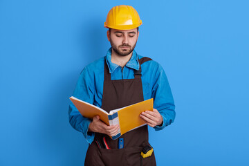 Architect going though paperwork, holding paper folder and reading something, looking down, wearing helmet, apron and blue uniform, posing isolated over color background.
