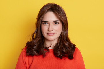 Fototapeta premium Attractive young Caucasian dark haired female posing against yellow background, dressed in casual orange sweater with calm face expression, looks at camera.