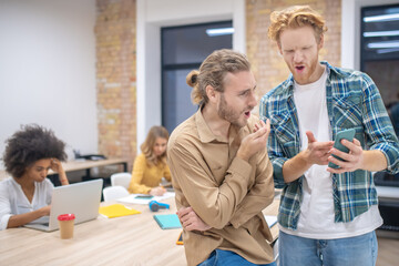Fototapeta premium Two male colleagues discussing something while looking at the phone abd looking enjoyed