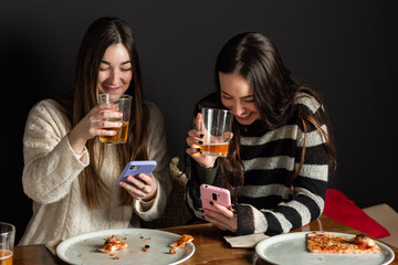 two girls drinking beer, using mobile phones and eating pizza.