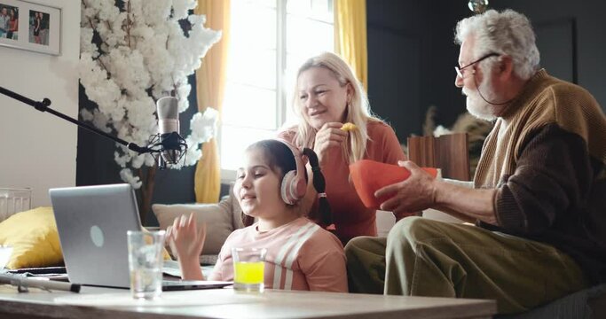 Little girl is streaming live at home using a laptop and a microphone. Grandma and grandpa eat popcorn and watch their granddaughter record a video blog