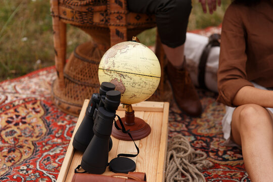 Eine Frau im Safari look sitzt draussen auf dem Tepich und  liest ein altes Buch, auf der Holzkiste steht ein Globus und ein fernglas.Sie ist auf der Reise, liebt Abendteuer