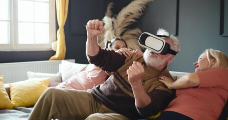 Grandpa uses VR for virtual car driving, grandma and granddaughter laugh. Happy family uses VR glasses - Powered by Adobe