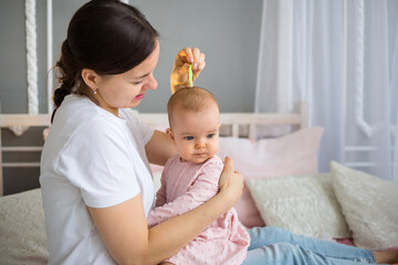 brunette mom combs her baby daughter on the bed in the room