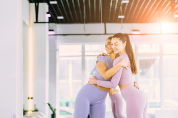 A team of two young athletic girls hold hands in front of the white interior of a loft studio. Female companions in the gym are resting after fitness, indoors, sun glare effect.