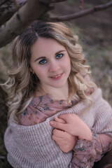 Young beautiful curly blonde woman in brown pleated skirt, pink blouse, covered shoulders with knitted scarf, sits in blooming peach sunset gardens. Girl enjoying walk outside the city, natural beauty